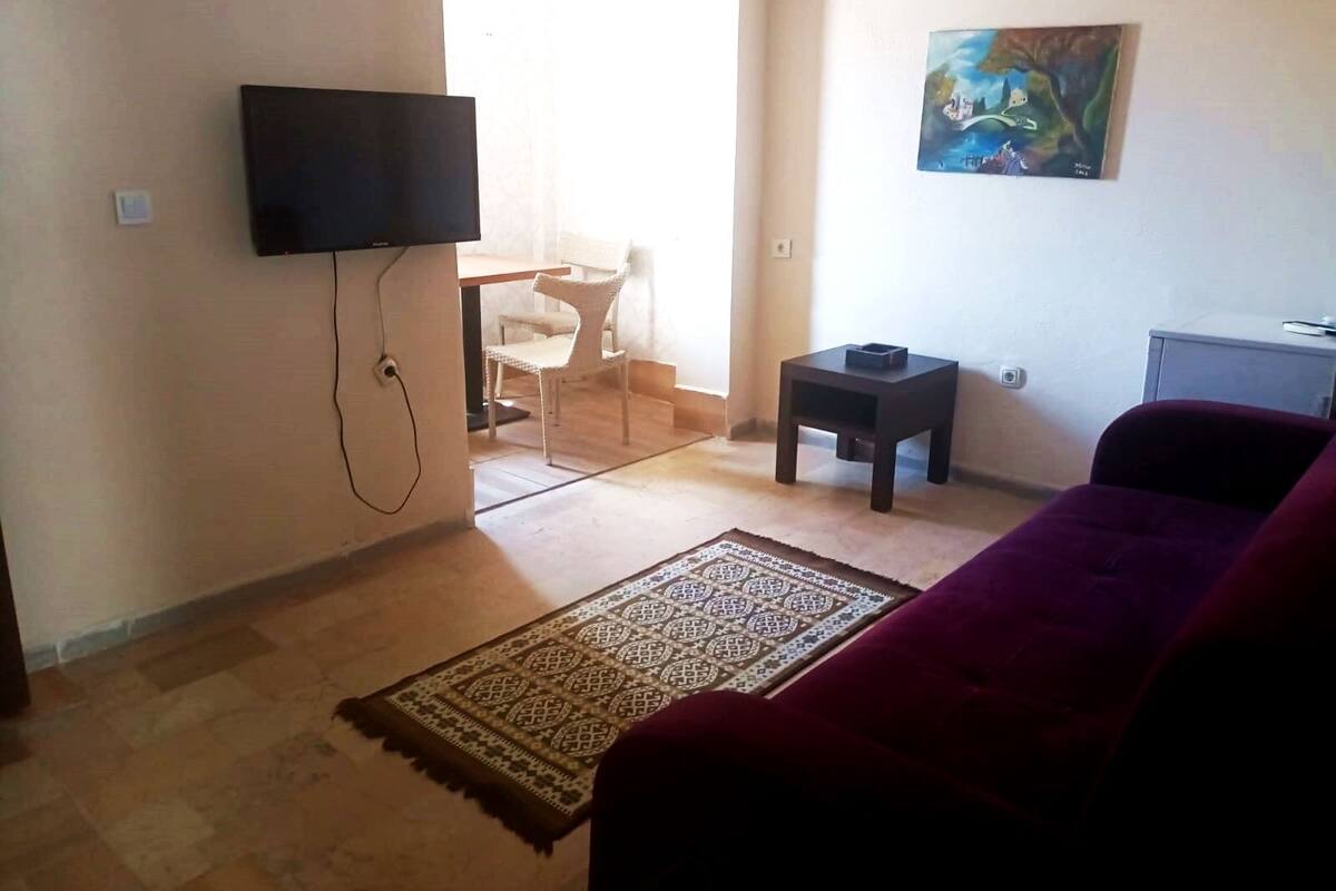 A living room with a flat-screen TV mounted on the wall, a small table, a chair, a sofa, and a rug on the floor.