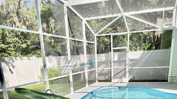 Ein Swimmingpool mit weißem Deck, umgeben von einer überdachten Liegefläche mit Stühlen und einem Blumentopf.