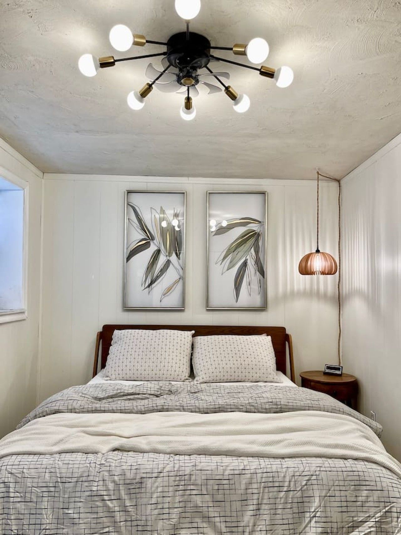 A bedroom with a bed, two wall-mounted artworks, a ceiling fan, and a bedside lamp.