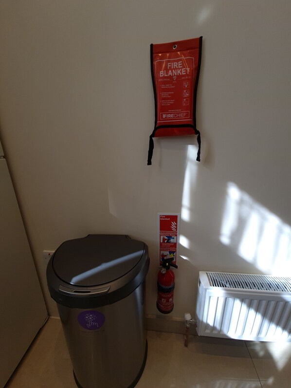 A kitchen with a trash bin, a fire blanket, a fire extinguisher, and a radiator.