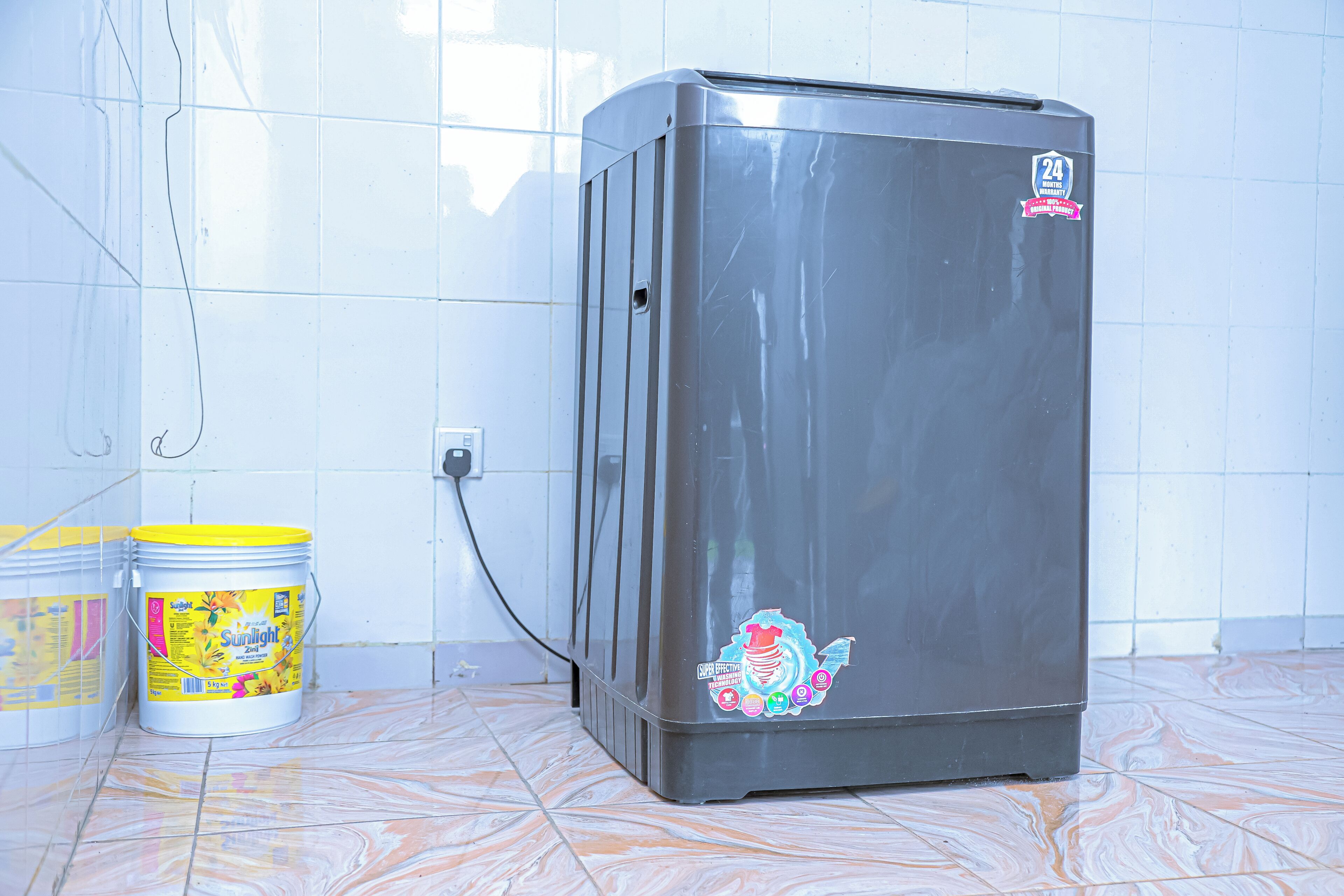 A compact refrigerator with a sticker, a power outlet, and two buckets on a tiled floor.