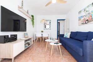 Un salon moderne dotĂ© dâun tĂ©lĂ©viseur Ă Ă©cran plat, dâun canapĂ© bleu, dâun coin repas avec une table et des chaises blanches, et dâun ventilateur de plafond.