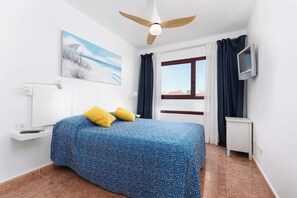 A bedroom with a bed, a ceiling fan, a TV, a window, and a painting on the wall.