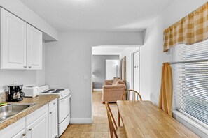 A compact kitchen with white cabinets, a stove, and a coffee maker. There is a dining table with chairs and a window with blinds. - The Charter House (Hollywood)