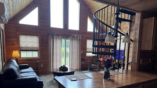 A spacious living area with a wooden ceiling, a black leather sofa, a dining table with chairs, a kitchen island with a vase of flowers, and a spiral staircase. - Rustic Elegance, 1 bedroom cabin in the heart of Jonesborough. (Jonesborough)
