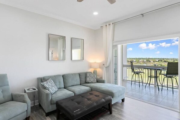 A modern living room with a grey sofa, a black ottoman, and a table with chairs. There are two mirrors on the wall and a window with a view of a green landscape. - Amazing Golf View Condo Best view in Reunion! (Kissimmee)