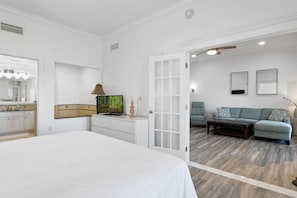 Ein Schlafzimmer mit Bett, Fernseher, Kommode und einer Tür zu einem Wohnbereich mit Sofa und Couchtisch.