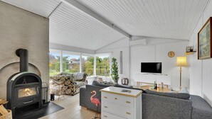 A modern living room with a wood-burning stove, a sofa, a TV, and a large window offering a view of the outdoors. - Cozy home in Karrebæksminde with WiFi (Karrebæksminde)
