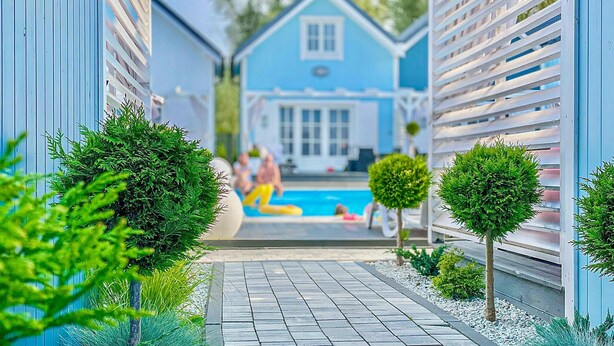 Ein schmaler Pfad führt zu einem Poolbereich, im Hintergrund ein blaues Haus.