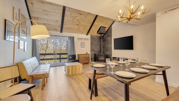 Un espace repas moderne avec une table en bois dressée pour six personnes, un canapé et une grande fenêtre donnant sur un paysage arboré.