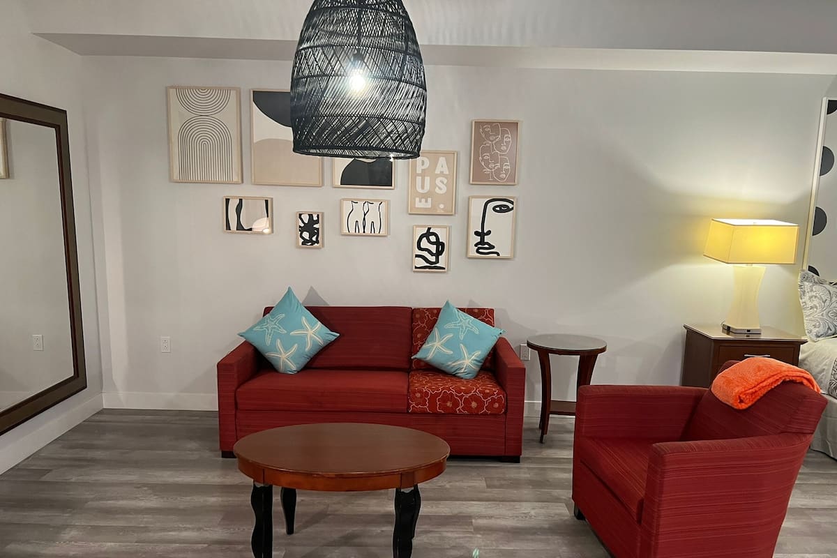 Un salon avec un canapé rouge, deux fauteuils assortis, une table basse en bois et une suspension noire. Les murs sont ornés d’œuvres d’art encadrées et une lampe est posée sur une table d’appoint.