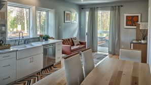 A modern kitchen with white cabinets, a stainless steel dishwasher, and a large window. There is a dining area with a wooden table and chairs, and a brown leather sofa. -  StaysPro - Hollywood Homes Minutes to Everything (Los Angeles)