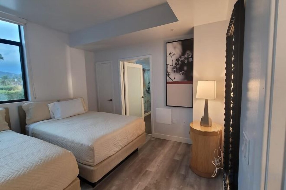 A hotel room with two beds, a bedside table with a lamp, a door leading to a bathroom, and a window with a view.