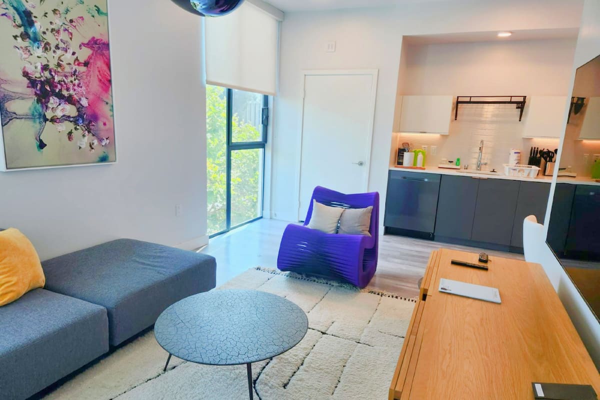 Un salon moderne avec un canapé gris, une petite table ronde et un fauteuil violet. On y trouve également un coin cuisine avec des placards gris et un plan de travail blanc.