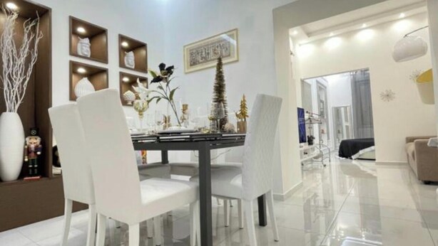 A modern dining area with a white table and chairs, a chandelier, and decorative items on shelves. - Nice and elegant apartment in the center (Oliveri)