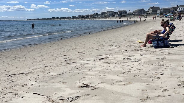 Una playa con cielo despejado, gente sentada en sillas de playa y algunas personas nadando en el agua.