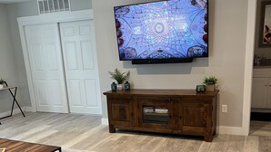 Una sala de estar con un televisor de pantalla plana grande montado en la pared, un mueble de madera para entretenimiento, un sofá verde y una alfombra estampada.