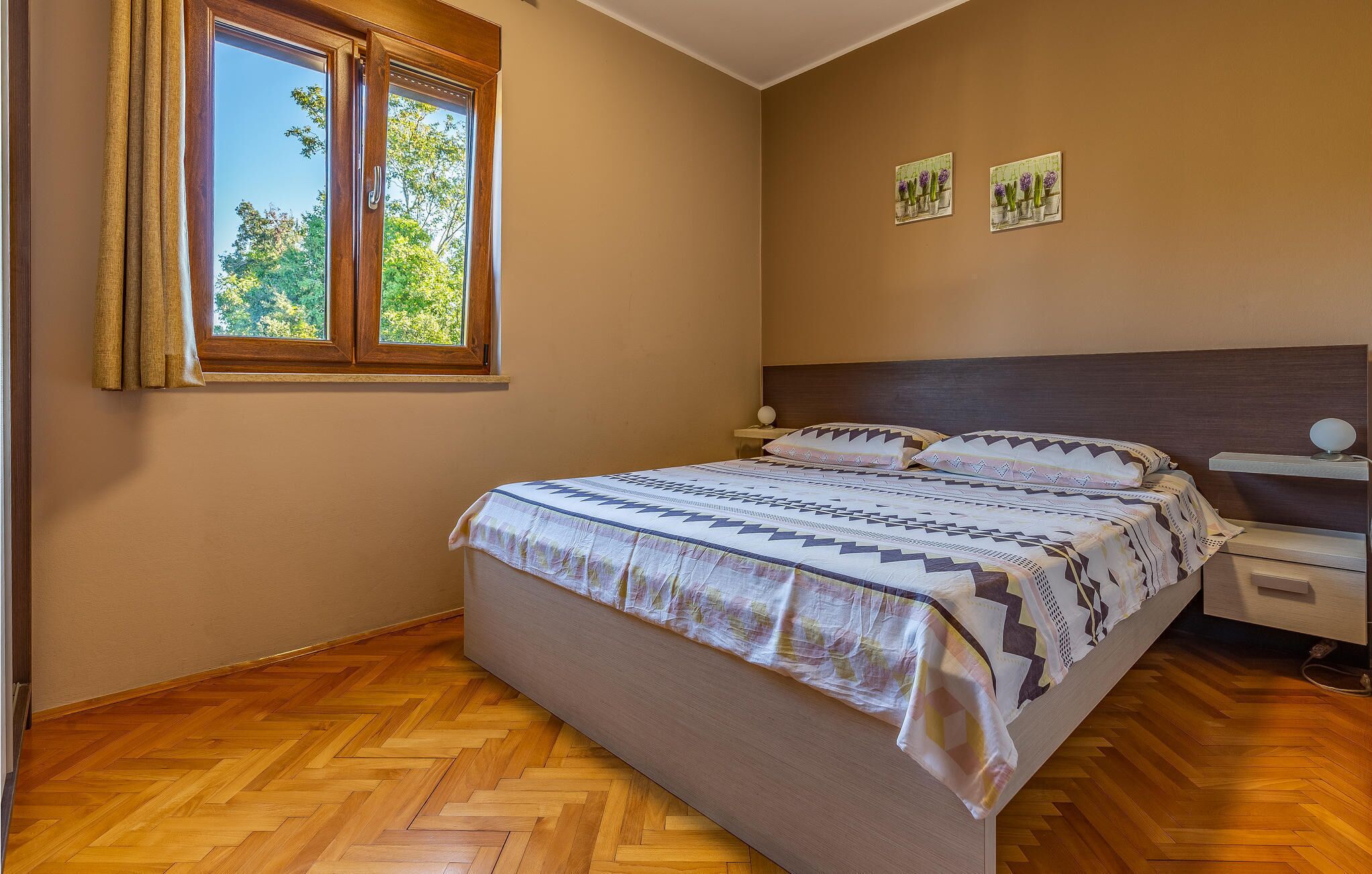 A bedroom with a wooden herringbone floor, a bed with patterned bedding, a nightstand with a lamp, and two wall-mounted paintings.