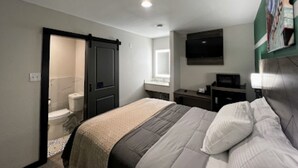A bedroom with a bed, a TV mounted on the wall, a bathroom visible through an open door, and a window providing natural light. - Bishop Arts Suites (Dallas)