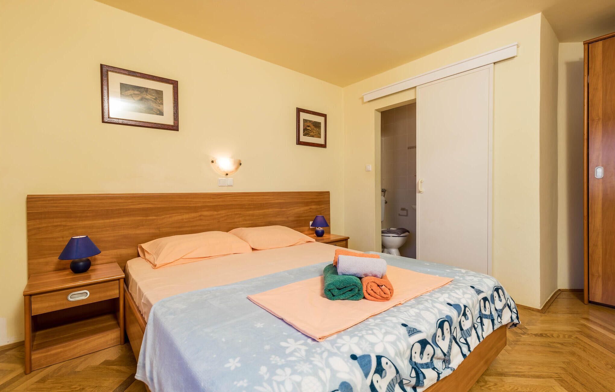 A bedroom with a wooden headboard, two bedside tables with lamps, a bed with a blue and white penguin-themed duvet, and two framed pictures on the wall.