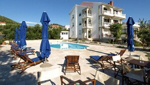 Ein Poolbereich mit Liegestühlen und Sonnenschirmen, ein Schwimmbecken und im Hintergrund ein mehrstöckiges Gebäude mit Balkonen.