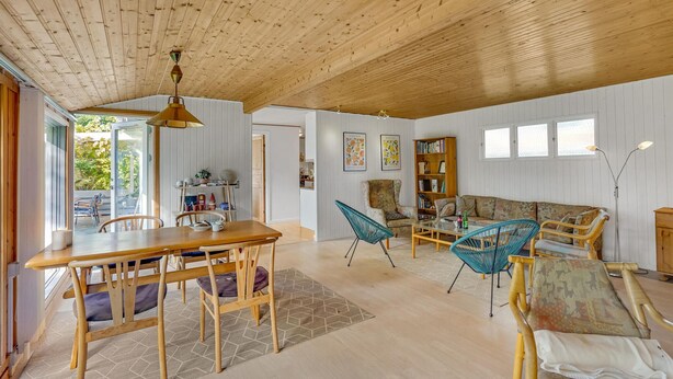 A spacious living room with a wooden ceiling, a dining area with a wooden table and chairs, and a sofa with patterned cushions.