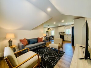 A modern living room with a sofa, chairs, a coffee table, and a black and white patterned rug. There is a dining area with a table and chairs, and a kitchen in the background.