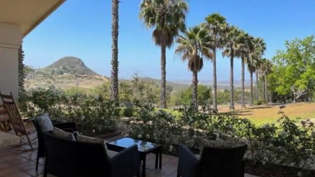 An outdoor seating area with chairs and a table, surrounded by plants and palm trees, overlooking a hilly landscape. - Ocean Breezes VRBO (Fallbrook)
