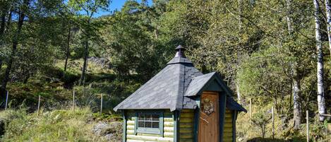 Uma pequena cabana de madeira com telhado inclinado e chaminé, rodeada por uma floresta densa.