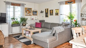 A cozy living room with a grey sofa, a wooden coffee table, and a television on a stand. There are potted plants and framed artwork on the walls.