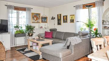 A cozy living room with a grey sofa, a wooden coffee table, and a television on a stand. There are potted plants and framed artwork on the walls.