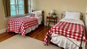 Two single beds with red and white checkered bedding, a wooden floor, a window with curtains, and a small bedside table with a lamp and a globe.