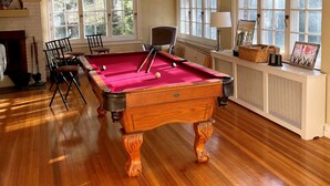 A pool table in a room with wooden floors, large windows, and a fireplace.