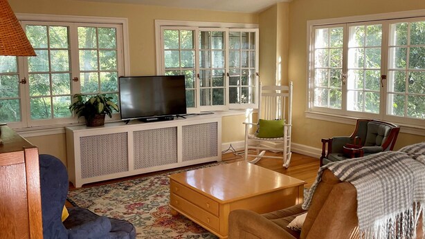 A spacious living room with a wooden floor, a large sofa, a coffee table, and a television on a white cabinet. There are multiple windows providing natural light and a view of greenery outside.