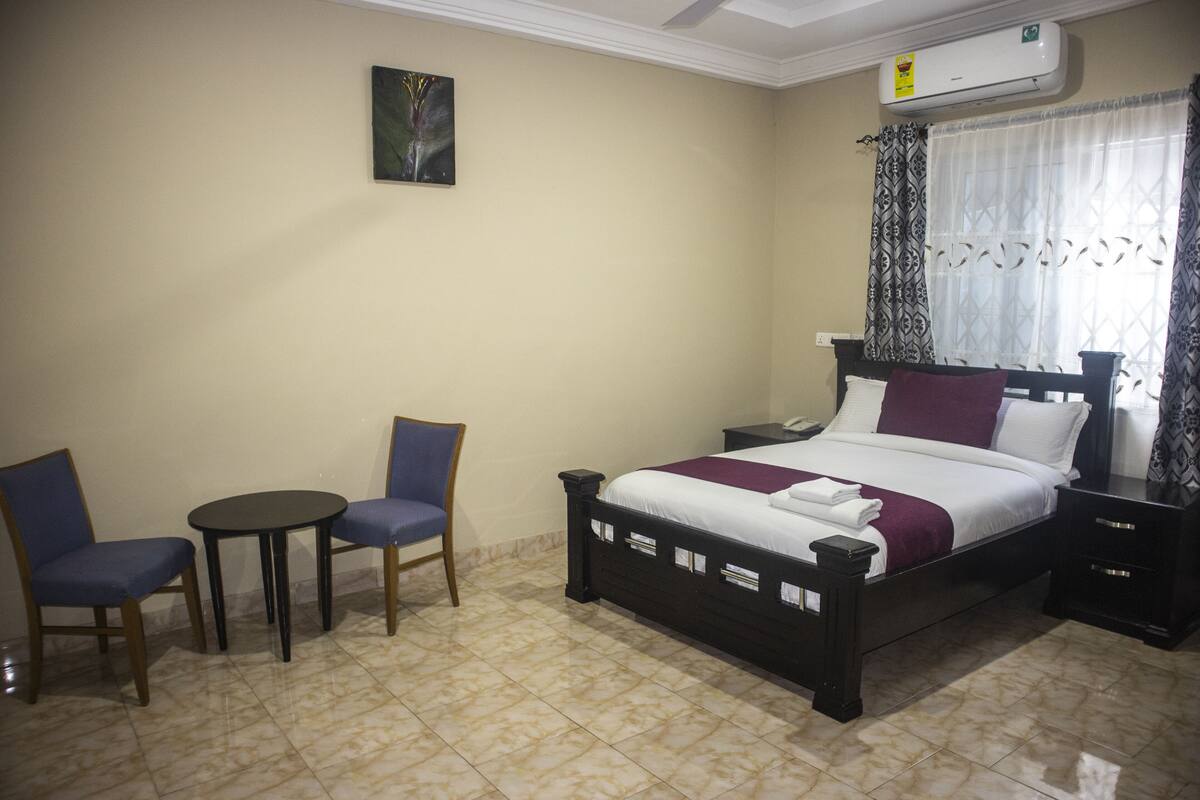 Habitación de hotel con una cama, dos sillas, una mesita pequeña, una mesita de noche y aire acondicionado instalado en la pared.