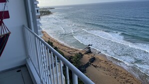 Un balcon avec vue sur l’océan, une plage et une côte lointaine parsemée de bâtiments.