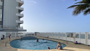 Un espace piscine entouré d’une clôture blanche, un bâtiment doté de balcons et un ciel dégagé.