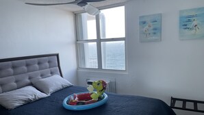 Une chambre à coucher comprenant un lit, un ventilateur de plafond, une fenêtre donnant sur la mer et deux tableaux accrochés au mur.