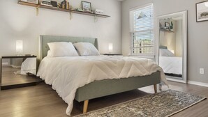 Un dormitorio con cama, mesitas de noche, un espejo, una ventana con persianas y una puerta que da a otra habitación.