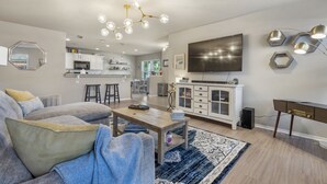 Una sala de estar moderna con un sofá, una mesa de centro y un televisor. Se observa una cocina al fondo con armarios blancos y un microondas.