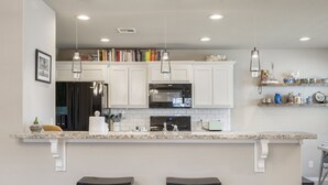 Una cocina con gabinetes blancos, microondas integrado, refrigerador de acero inoxidable y encimera de granito. Hay dos taburetes de bar y una lámpara colgante.
