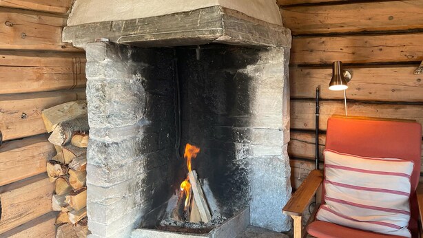 A stone fireplace with a burning fire, a stack of firewood to the left, and a red chair with a striped cushion to the right. - 3 bedroom nice home in Kvam (Kvam)