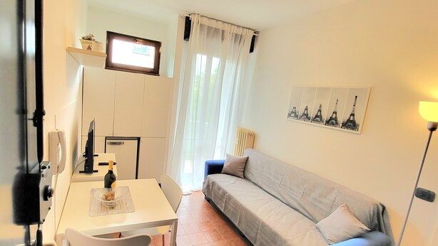 A small, well-lit room with a white table, a grey sofa, a TV mounted on the wall, and a framed picture of the Eiffel Tower. - Apartment in front of the hospital  (Modena)
