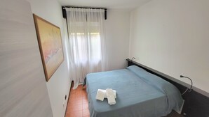 A small, clean room with a bed, a painting on the wall, and a window with sheer curtains. - Apartment in front of the hospital  (Modena)