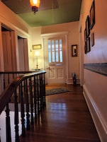 Un pasillo con suelo de madera, una escalera con barandilla oscura, una puerta con ventanal y una pared decorada con cuadros.
