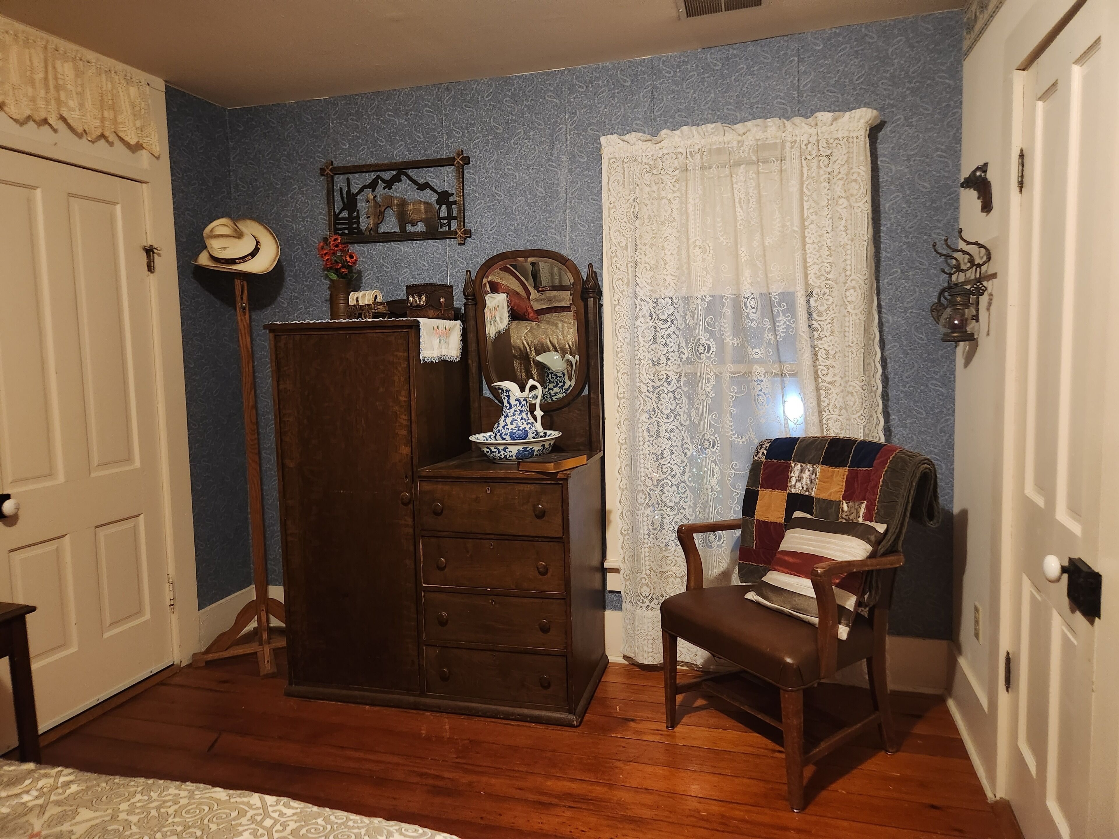 A room with a wooden dresser, a chair with a patchwork quilt, a mirror, and a decorative wall piece.