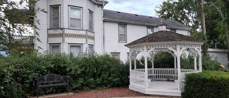 A white two-story house with a covered porch and a small garden area with a bench and a gazebo.