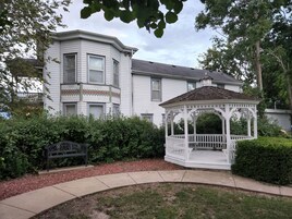 Una casa blanca de dos pisos con un porche cubierto y un pequeño jardín con un banco y un pabellón.