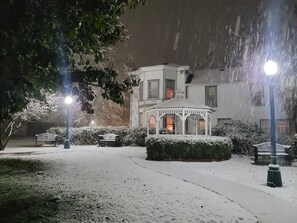 Um gazebo em um jardim coberto de neve, com bancos e um edifício ao fundo.