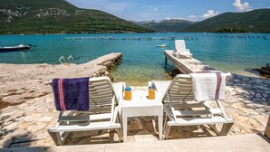 A seaside terrace with two white lounge chairs, a table, and a view of a calm lake with boats and mountains in the background.